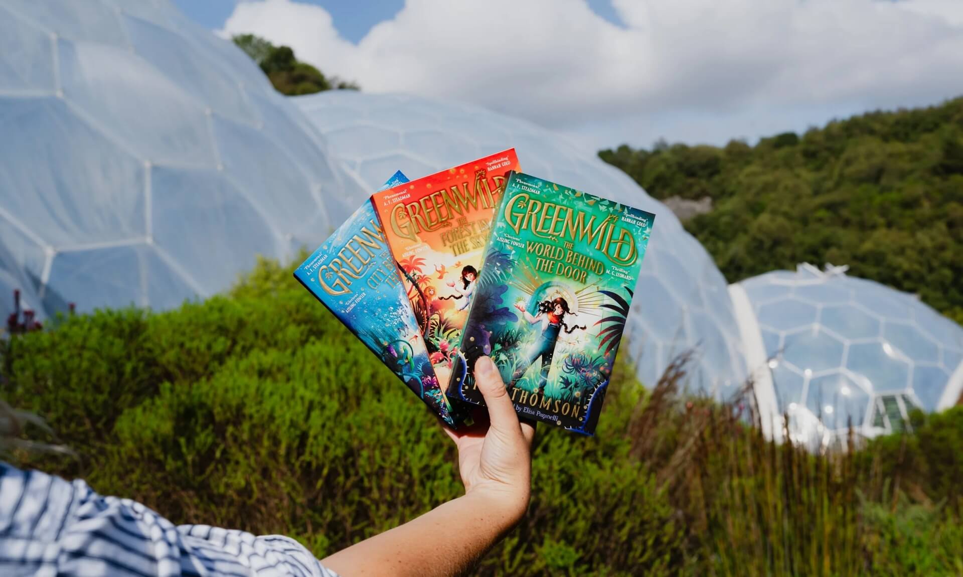 Hand holding the three Greenwild books in front of Eden Project's Biomes