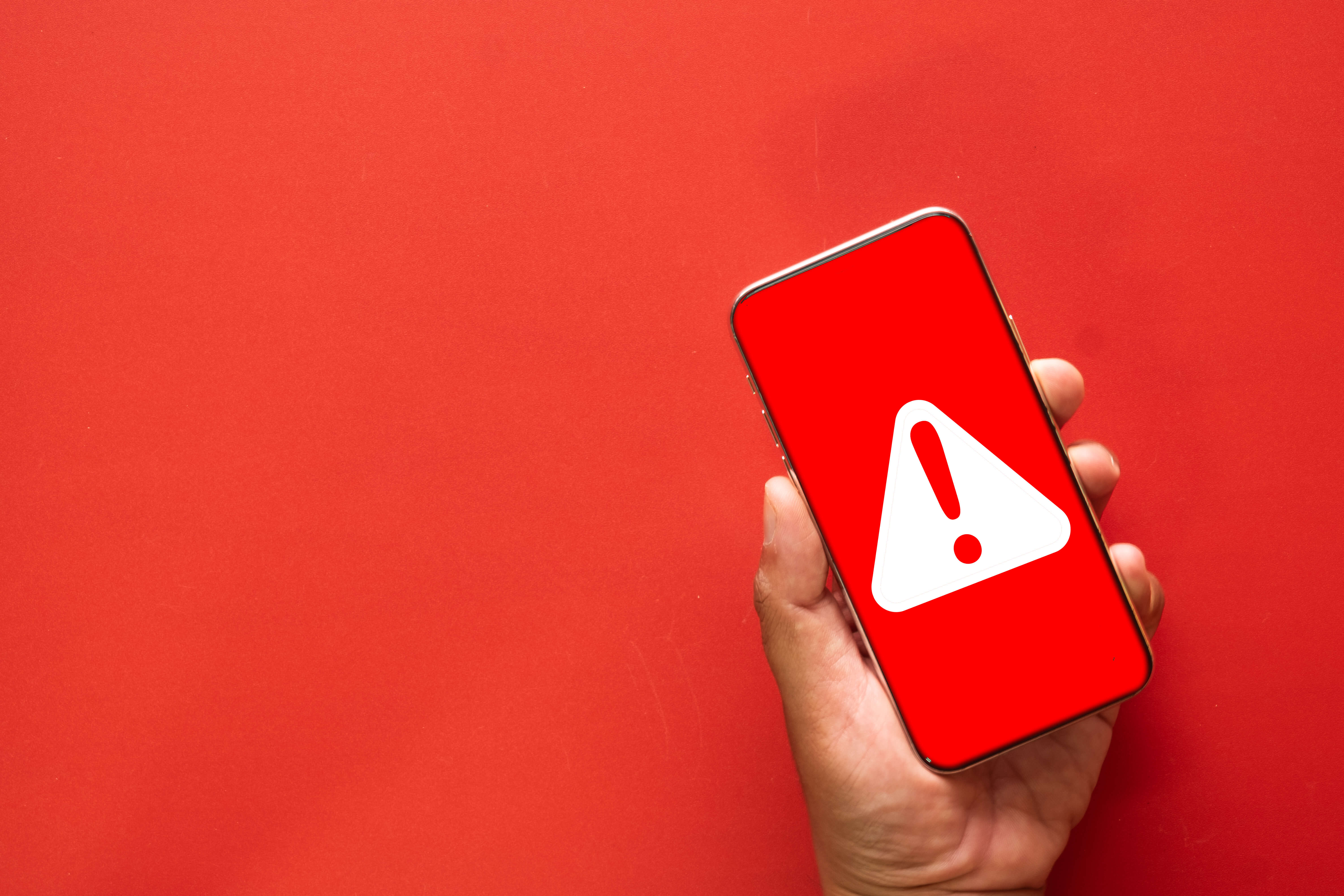 A hand holding a smartphone showing a red emergency alert screen with a white warning triangle