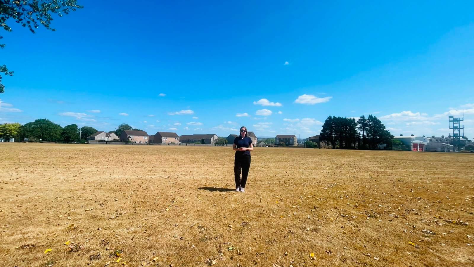 Anna Gelderd MP standing in a sun-parched field of straw-coloured grass in South East Cornwall under a clear blue sky