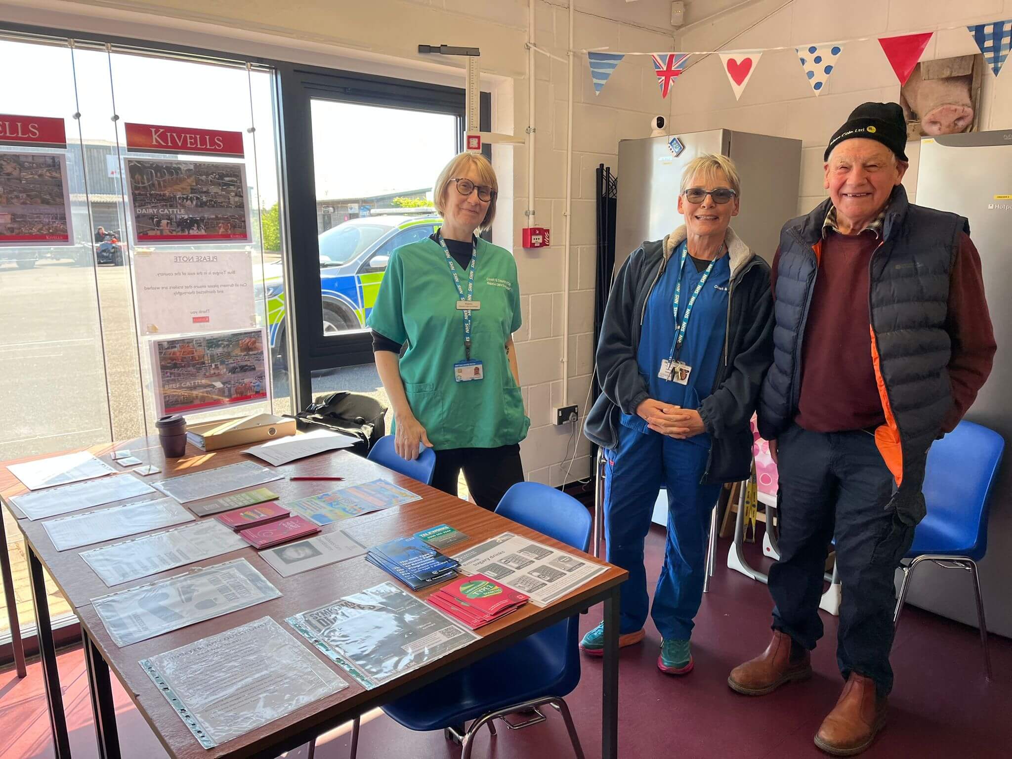 Two NHS staff and a farmer beside a table of health leaflets inside the Market Health Hub at Holsworthy livestock market