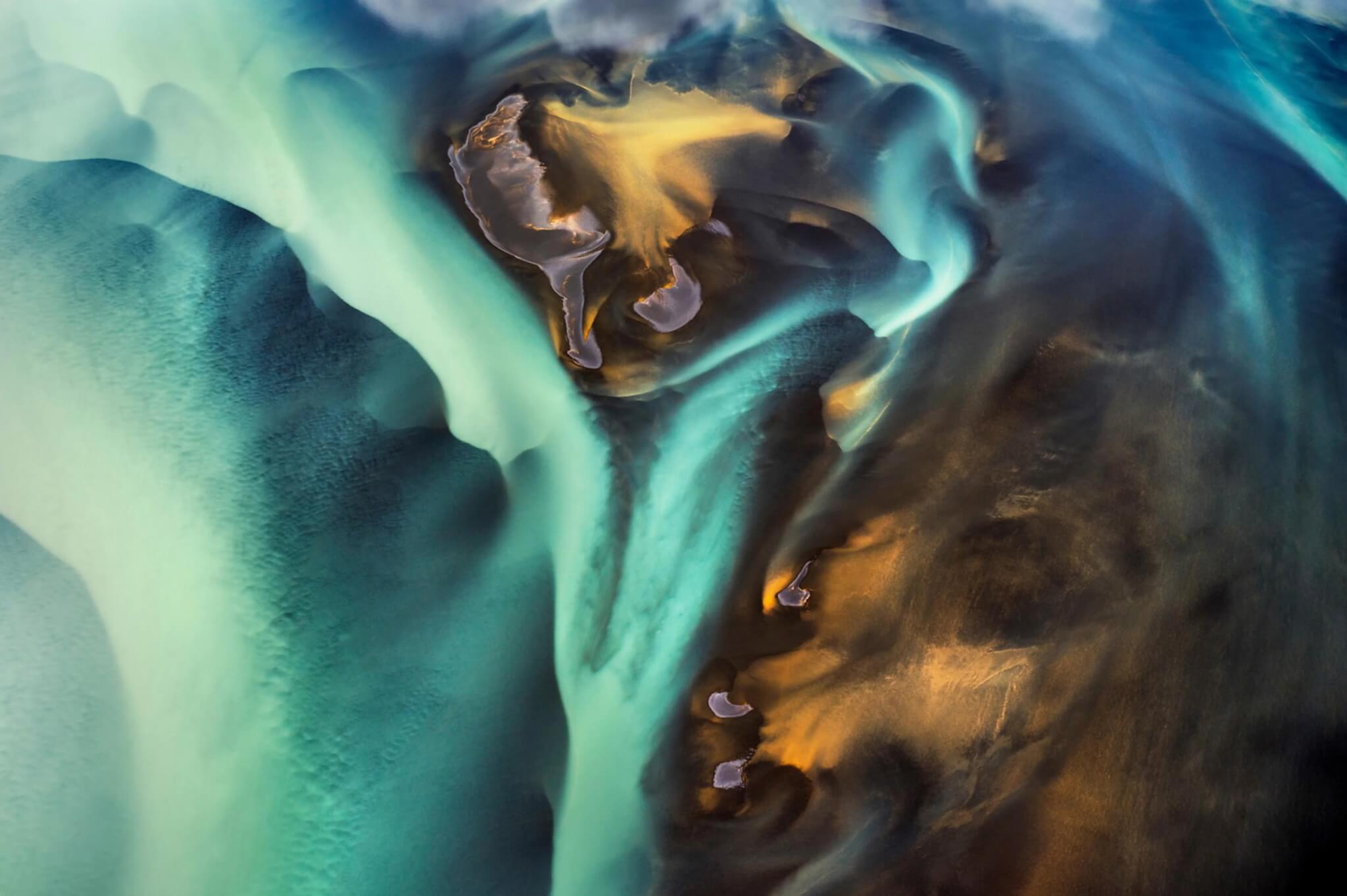 Aerial view of a glacial river in Iceland, turquoise and gold sediment swirling through dark volcanic sand