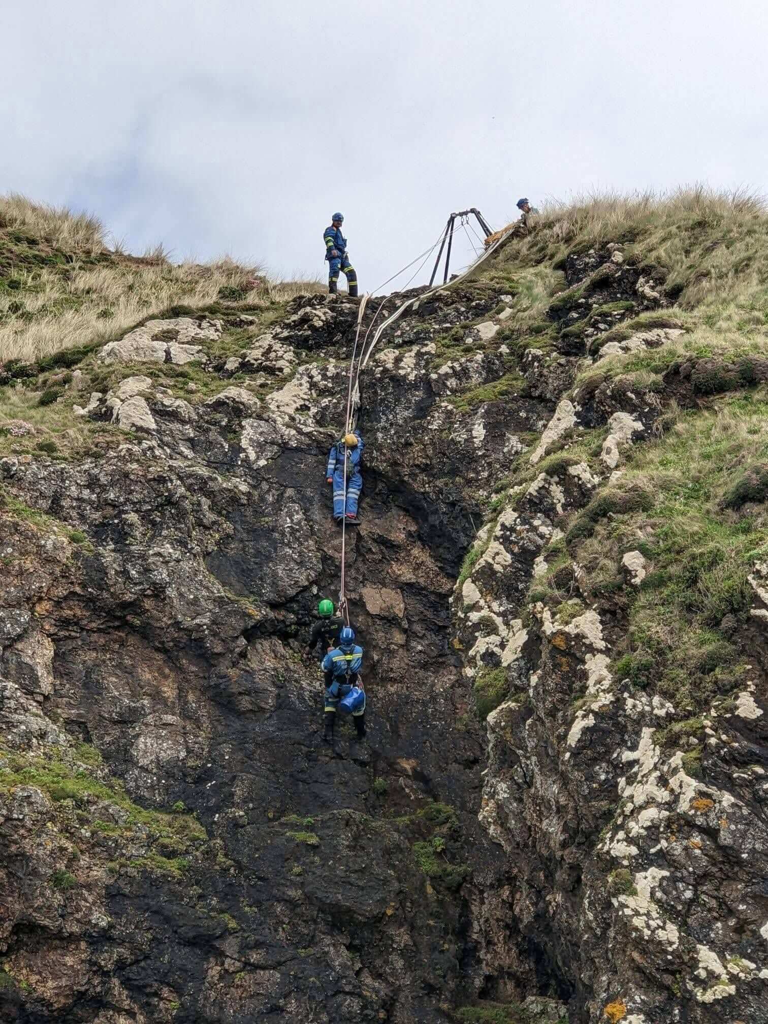 Coastguard rescue officers roped down a steep cliff face with a rope tripod rigged on the clifftop above