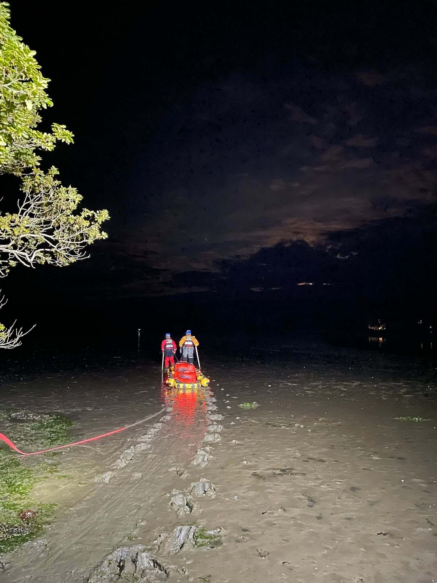 Two rescue officers pushing a mud rescue sled across an estuary at night, lit by floodlight
