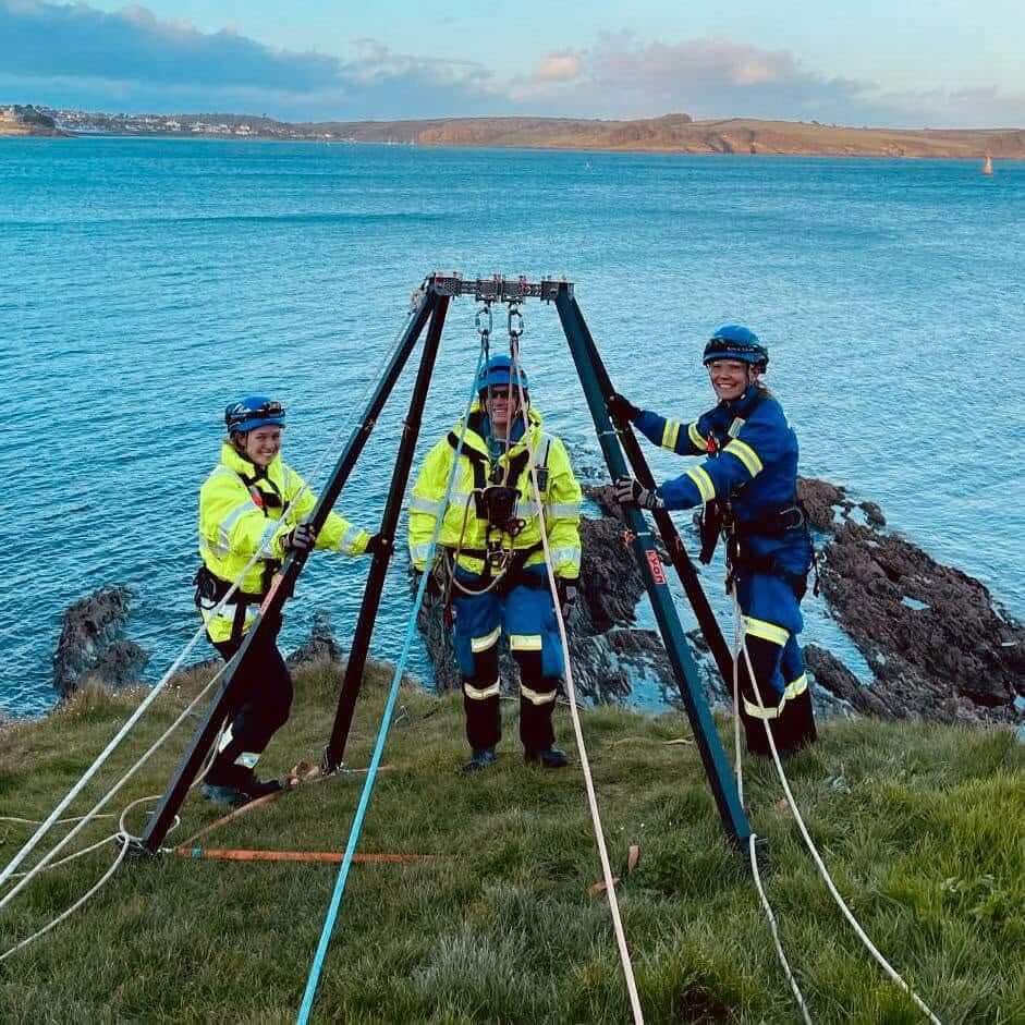 Three Coastguard rescue officers standing with a rope rescue tripod rigged on a grassy headland above the sea