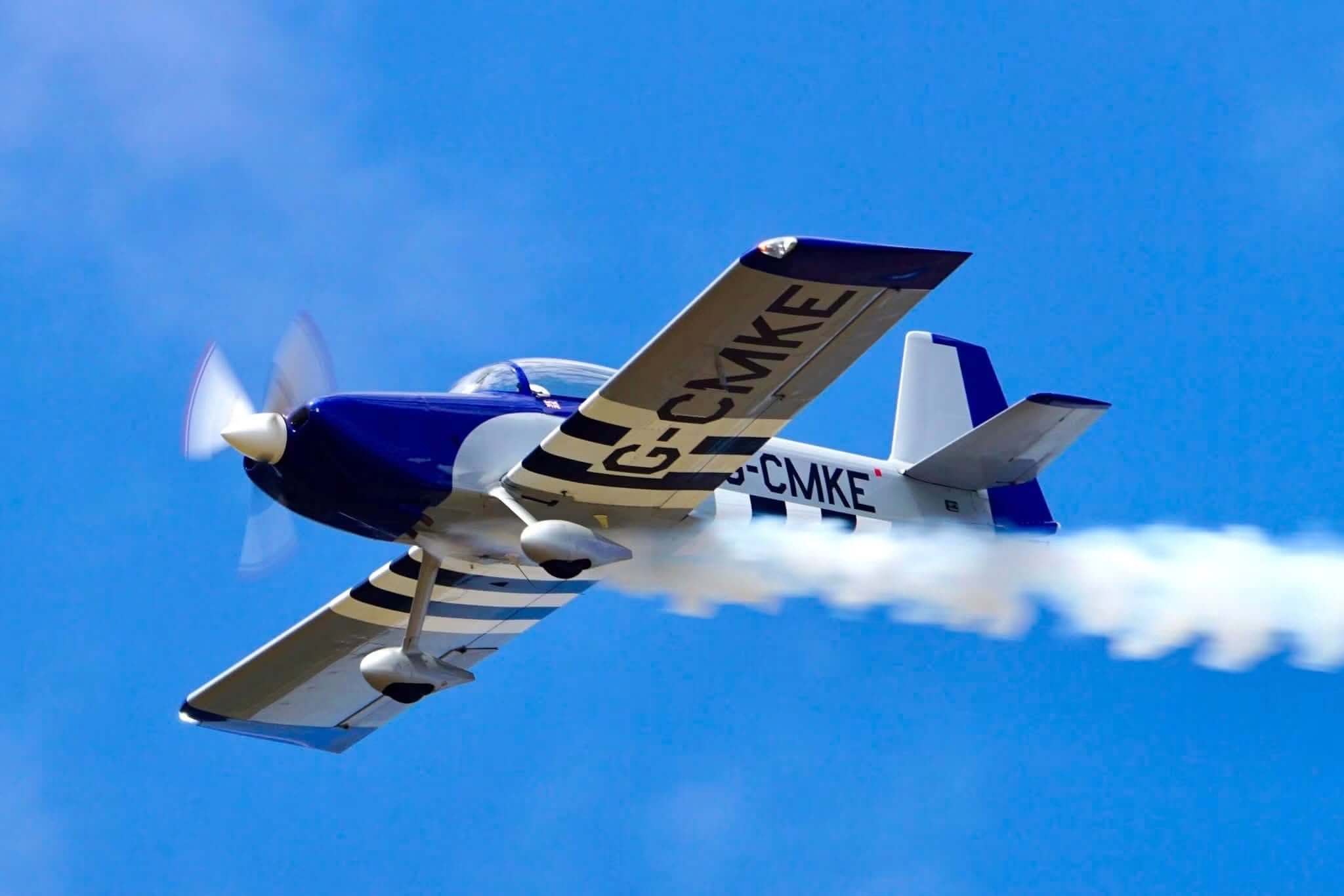 Blue and gold Van's RV aerobatic aircraft trailing white smoke against a clear blue sky