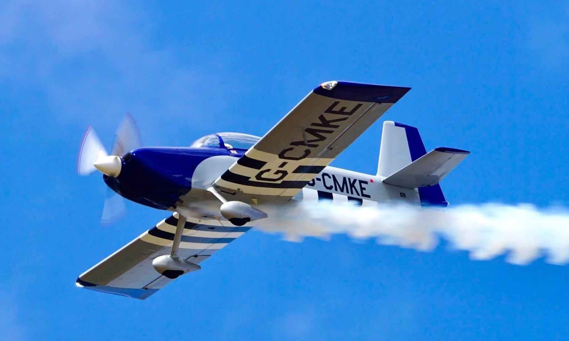 Blue and gold Van's RV aerobatic aircraft trailing white smoke against a clear blue sky