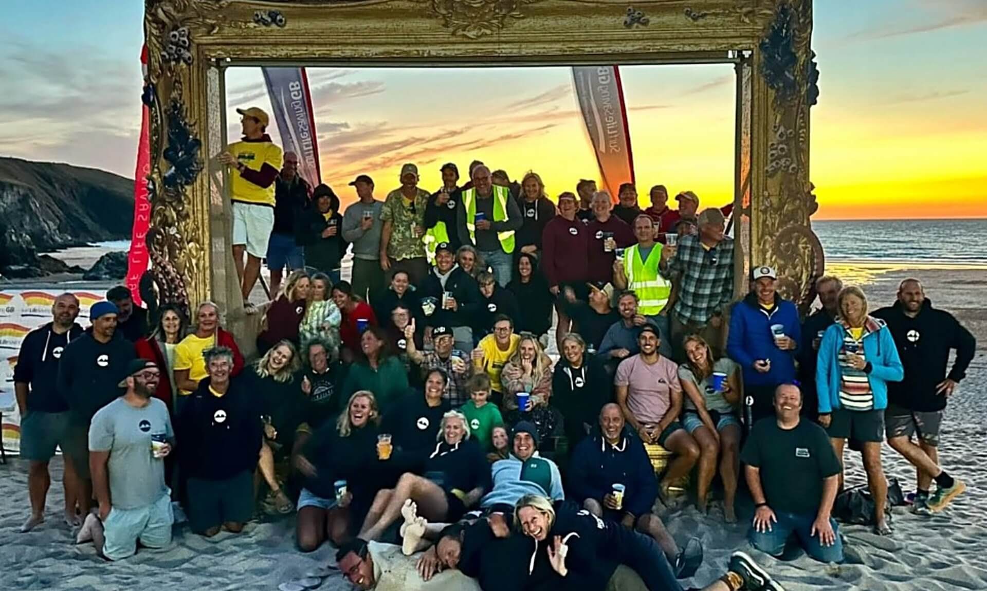 Volunteers and competitors gather under a large picture frame on Holywell Bay beach at sunset