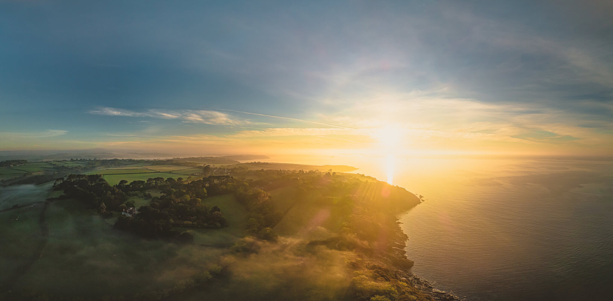 Aerial view of the Helford coast at sunrise, with a wooded headland, fields and the sea near Helford Passage