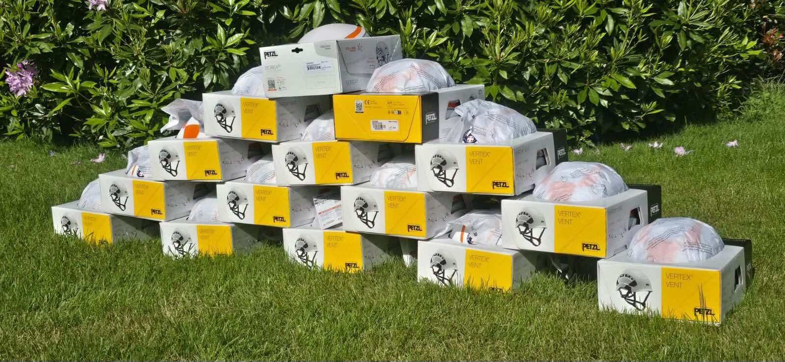 Dozens of boxed Petzl helmets stacked into a pyramid on grass in front of a hedge