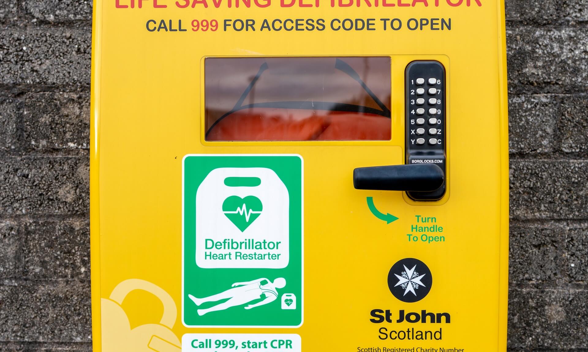 A yellow public access defibrillator cabinet mounted on a brick wall, reading Life Saving Defibrillator, Call 999 for access code to open.