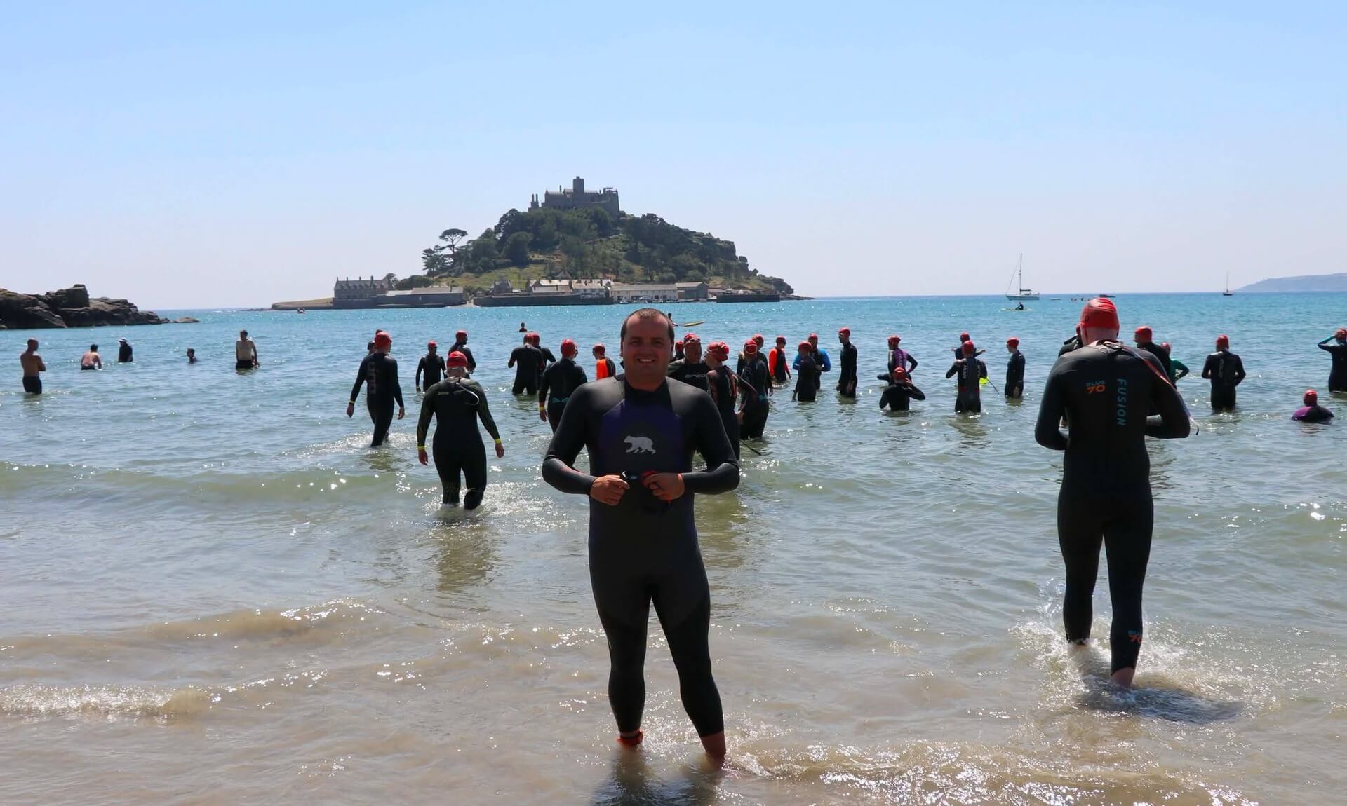 Matt Trewhella in a wetsuit on the beach at Marazion with St Michael's Mount behind him and swimmers gathering for a mass swim