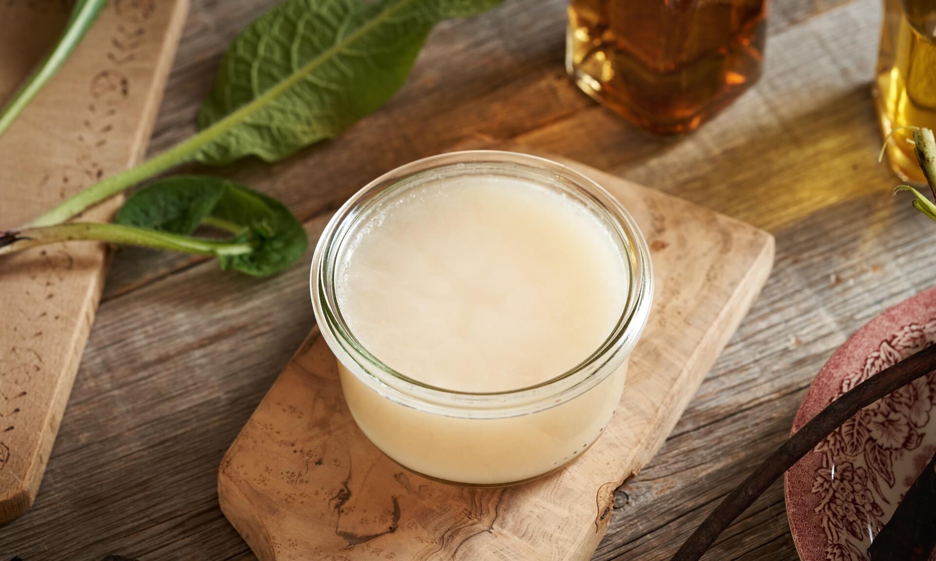 Homemade herbal skin balm in a glass jar on a wooden board with fresh herbs and infused oils