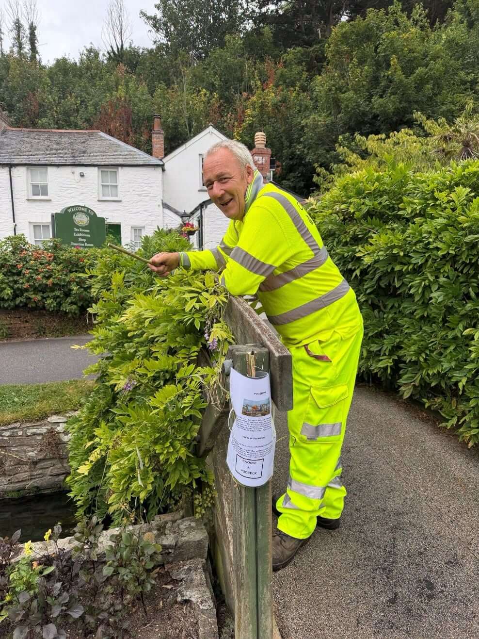A gardener in hi-vis reaching over the Wisteria Bridge at Trenance Gardens in Newquay, a Pooh sticks holder attached below