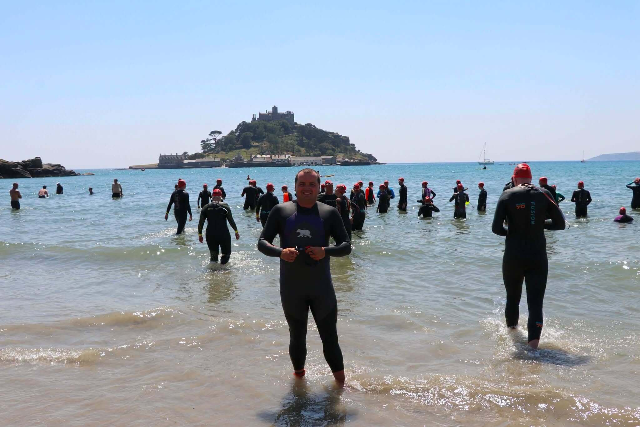 Matt Trewhella in a wetsuit on the beach at Marazion with St Michael's Mount behind him and swimmers gathering for a mass swim