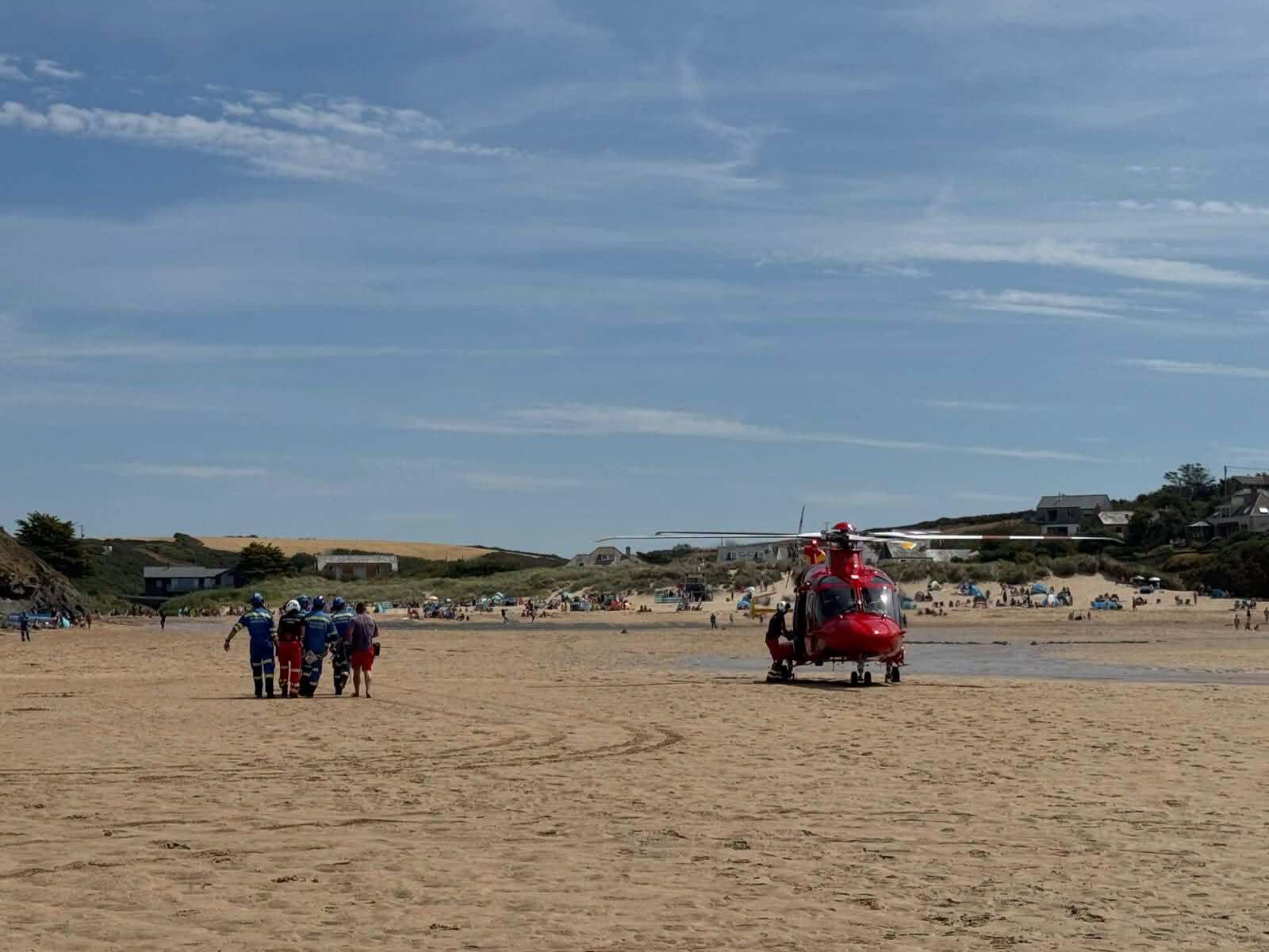 Cornwall Air Ambulance red helicopter on a busy Porthcothan beach as responders walk a casualty across the sand