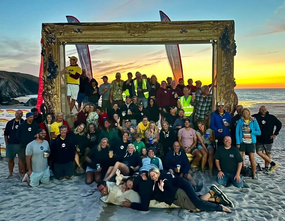 Volunteers and competitors gather under a large picture frame on Holywell Bay beach at sunset