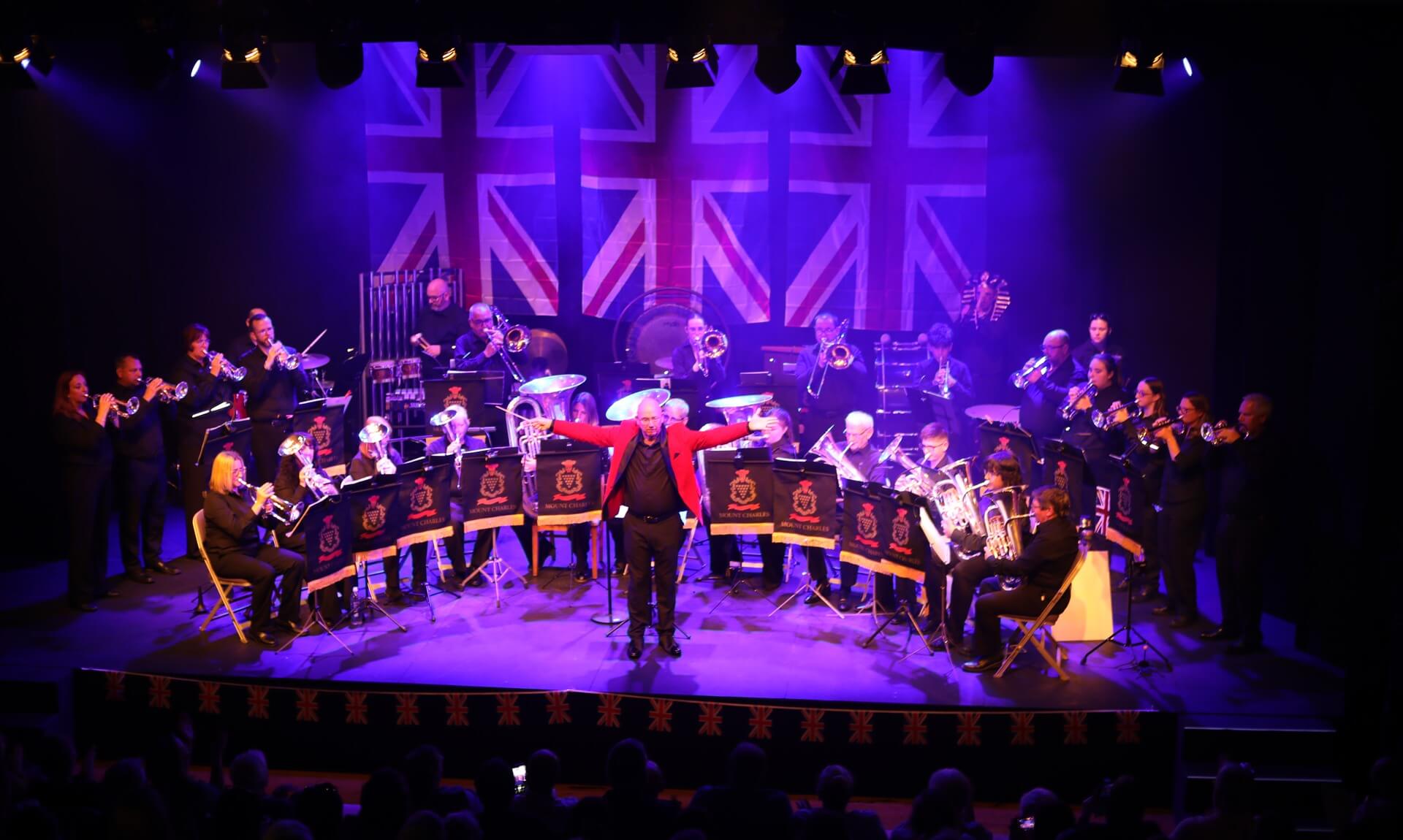 Mount Charles Band performing on stage at The Keay Theatre with a conductor in a red jacket and a Union Jack backdrop
