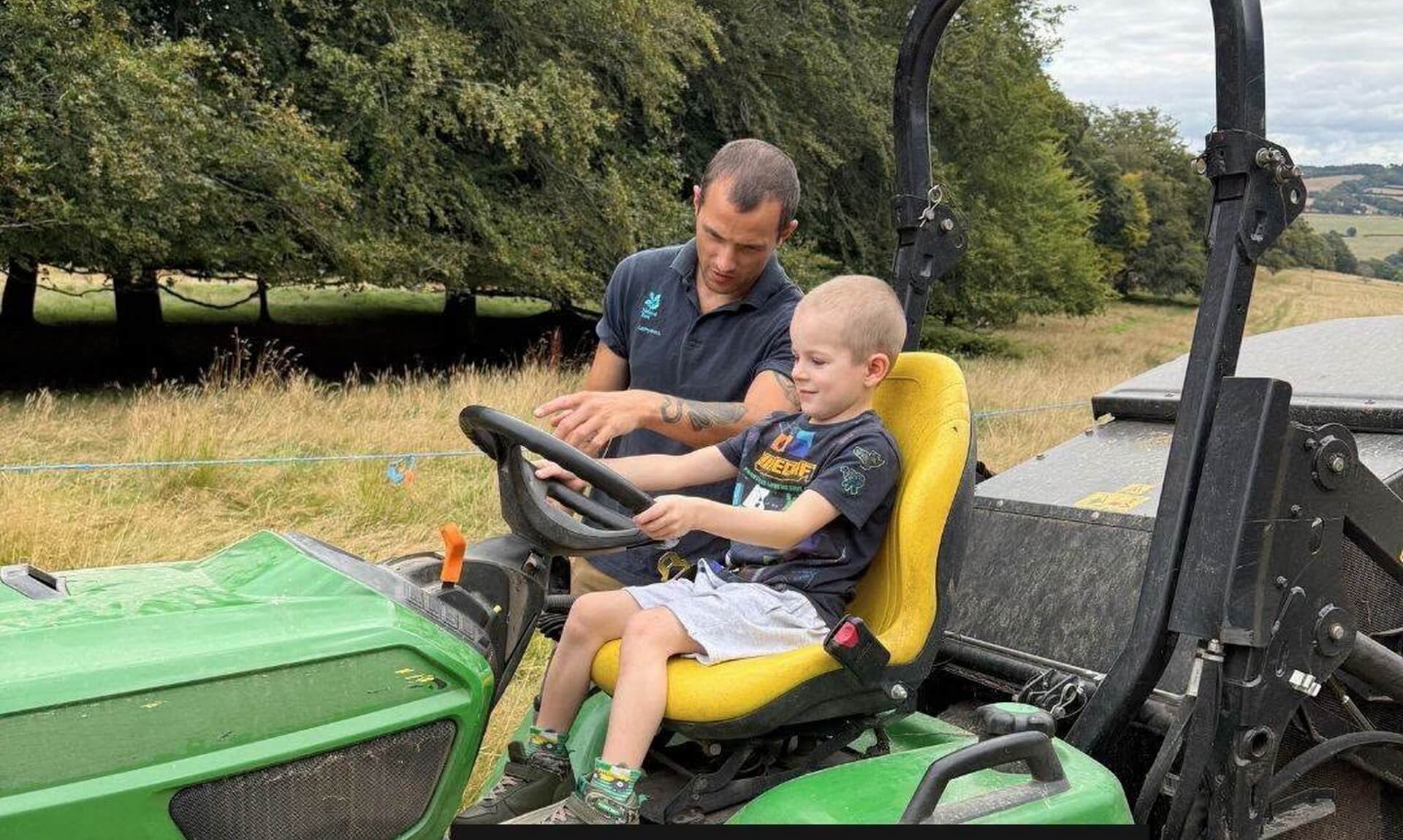 A National Trust ranger showing a young child how to steer a green ride-on mower in a field at Lanhydrock near Bodmin