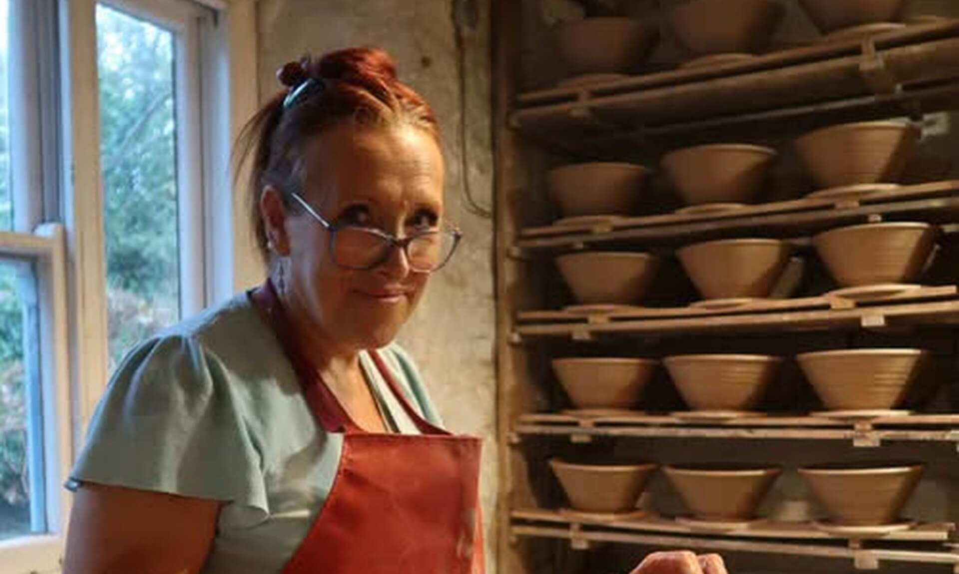 A potter in a red apron at the wheel at the Leach Pottery, with shelves of drying bowls behind her