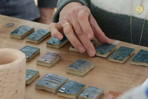 A hand arranging rows of blue glazed test tiles on a wooden bench
