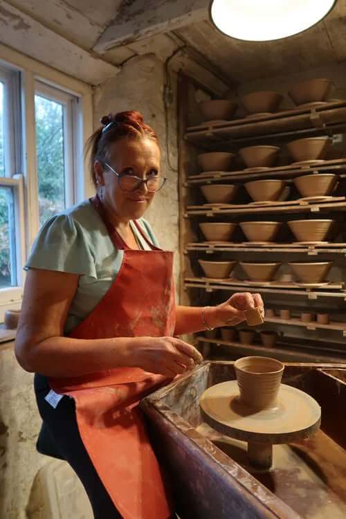 A potter in a red apron at the wheel at the Leach Pottery, with shelves of drying bowls behind her