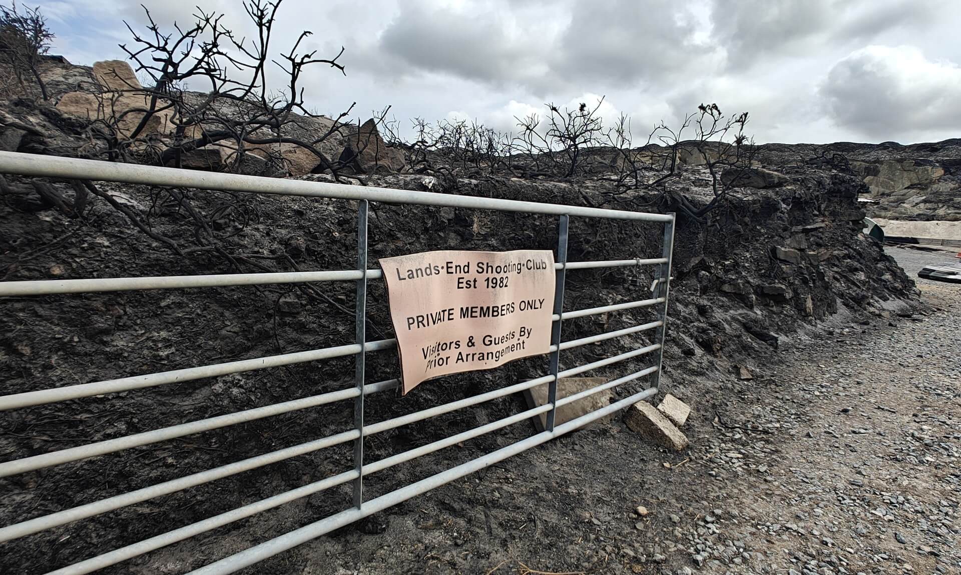 Burnt gate with Land's End Shooting Club sign amid wildfire-scorched ground