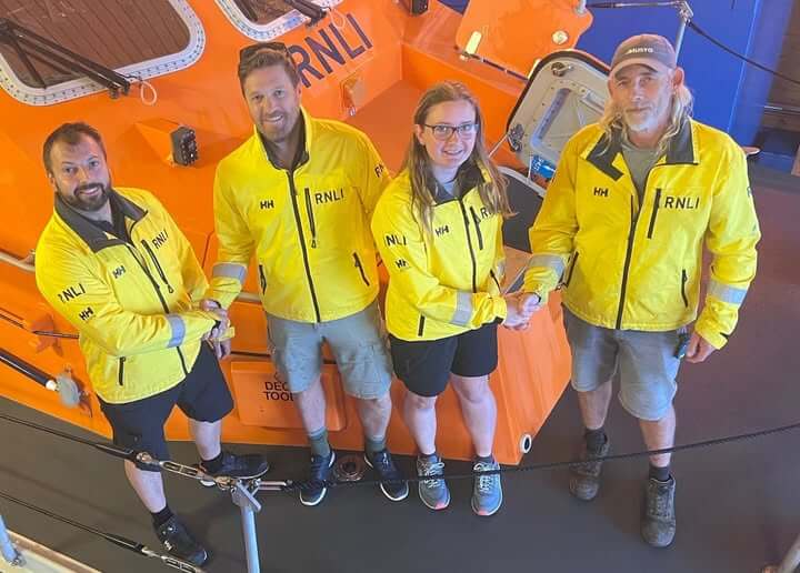 Four members of The Lizard RNLI crew in yellow kit standing beside the orange lifeboat in the boathouse