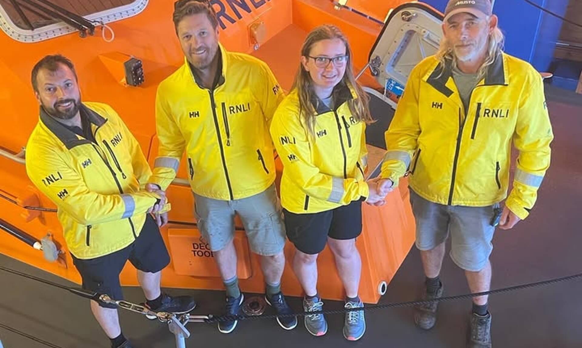Four members of The Lizard RNLI crew in yellow kit standing beside the orange lifeboat in the boathouse
