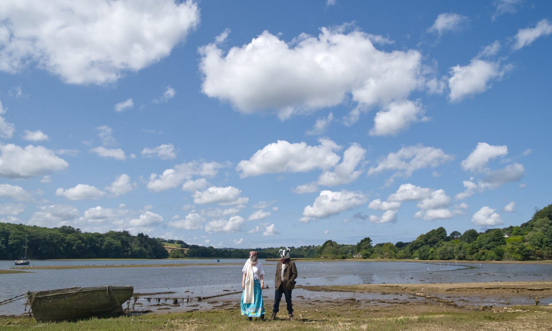 Meles Lutra in otter and badger masks on the shore of a Cornish creek under a big cloudy sky