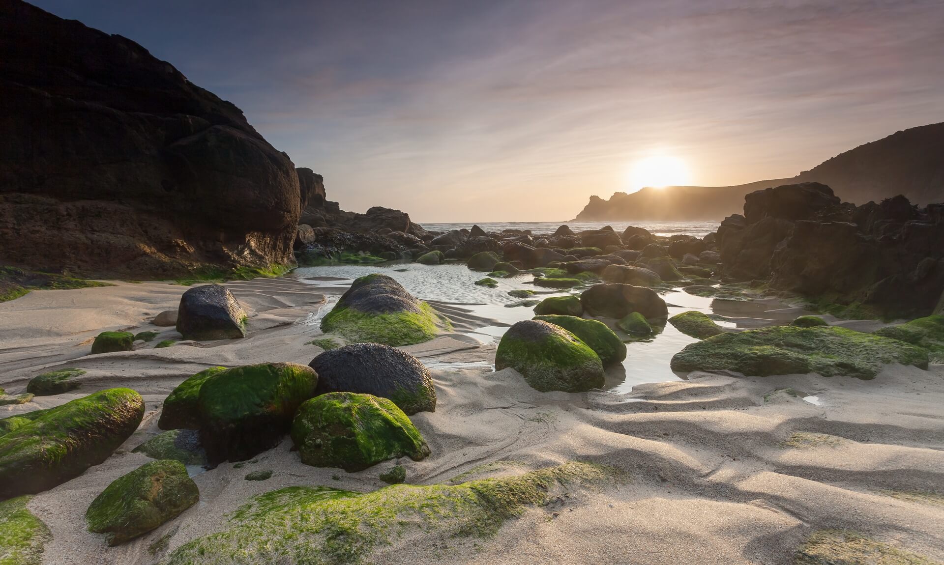Nanjizal beach near Land's End at low tide, moss-covered boulders and pools of water on golden sand at sunset