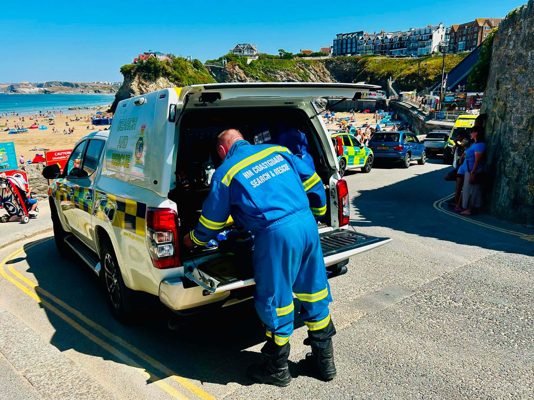HM Coastguard search and rescue vehicle parked above Towan Beach in Newquay, an officer at the open tailgate, an ambulance and sunbathers on the sand behind
