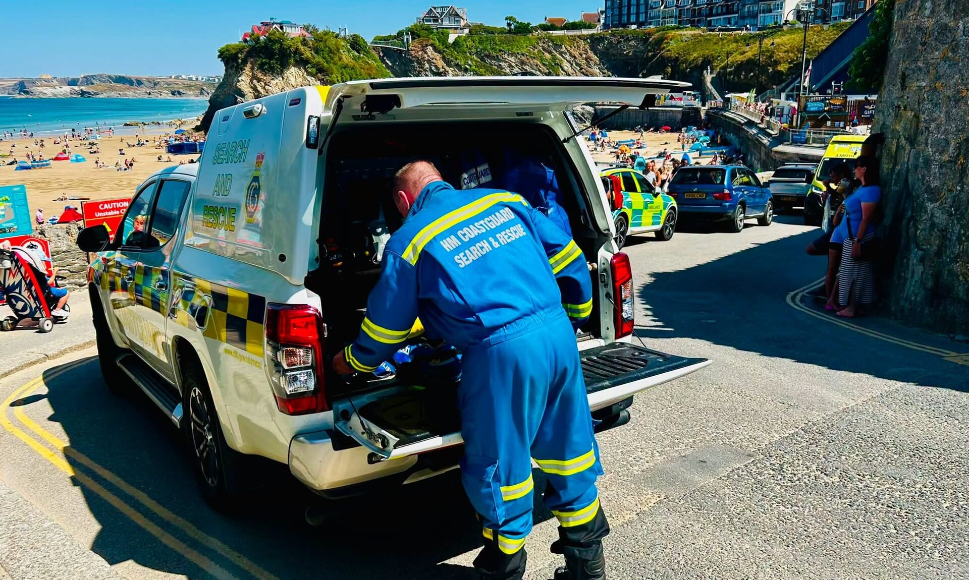 HM Coastguard search and rescue vehicle at the top of the ramp down to Towan Beach in Newquay, an officer working at the open tailgate with an ambulance and a busy beach behind