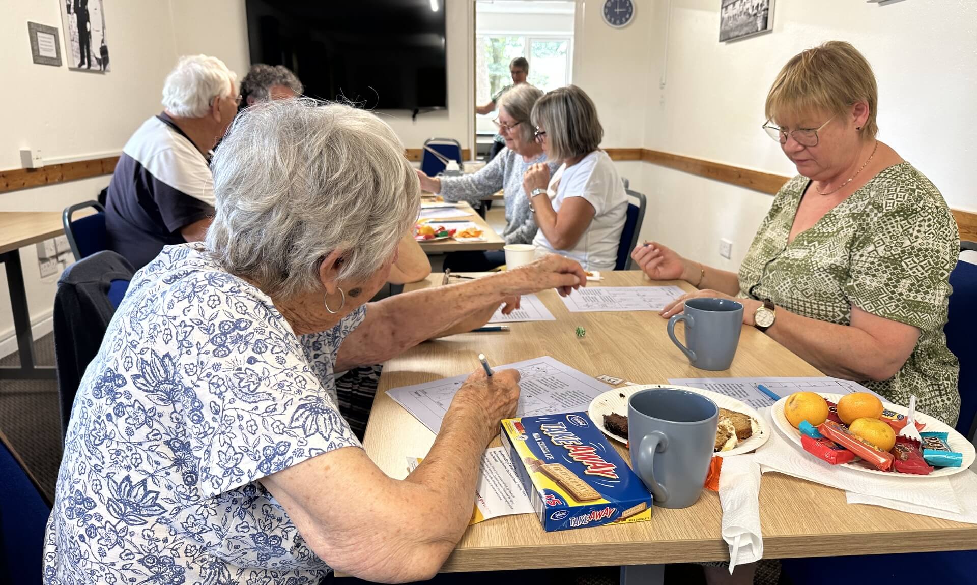Oddfellows members at a beetle drive around a table with puzzle sheets, mugs, biscuits and fruit at Kea Community Centre, Truro