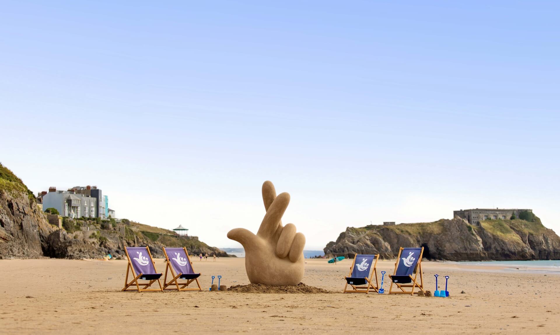 Giant crossed-fingers sand sculpture on a beach with National Lottery deckchairs, for The National Lottery Summer Hotspots