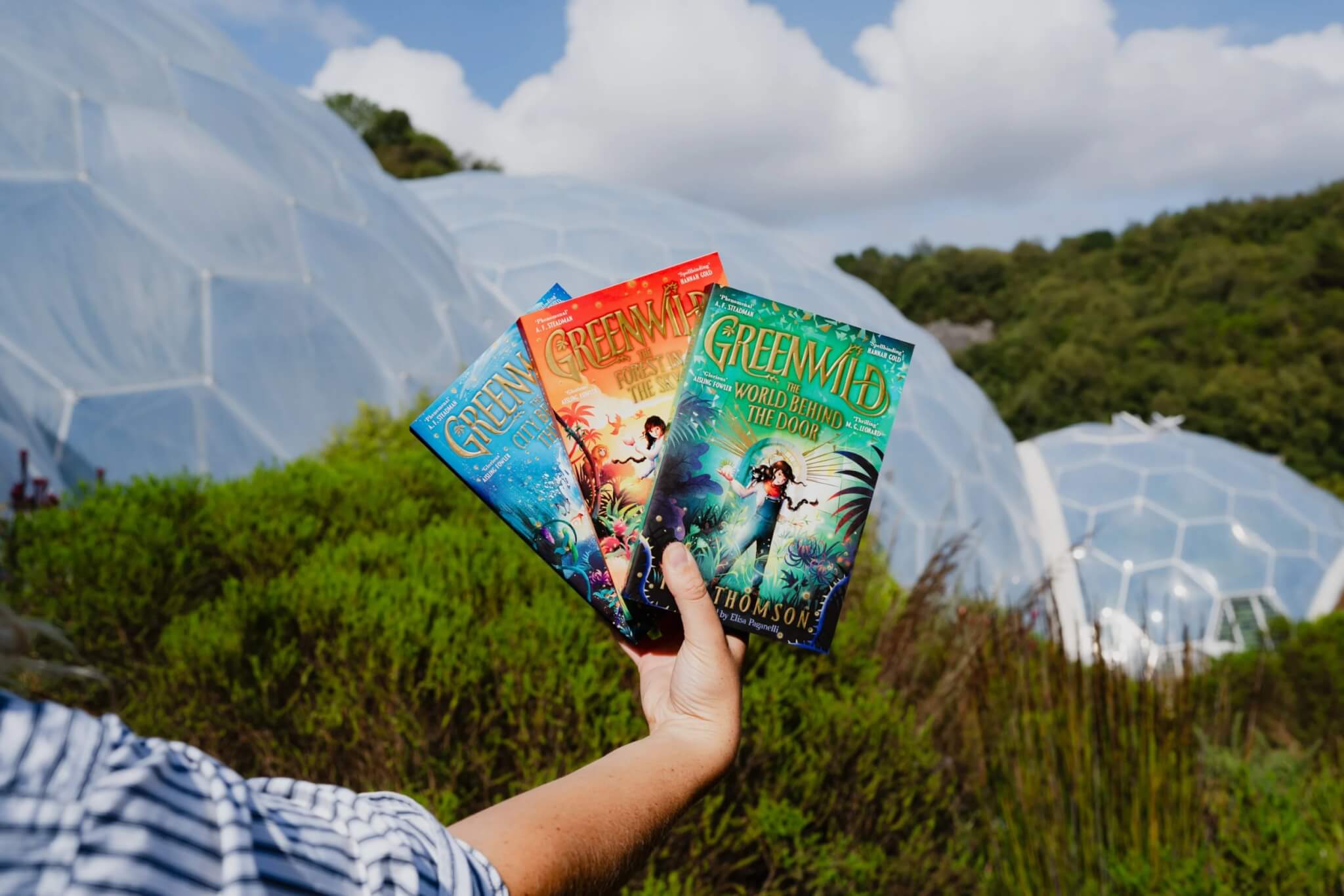 Hand holding the three Greenwild books in front of Eden Project's Biomes