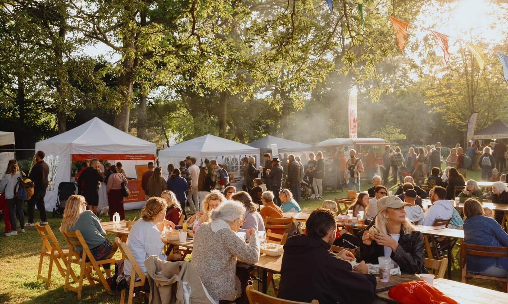 Crowds eating at long tables under trees hung with bunting at a Penzance food festival in golden evening light