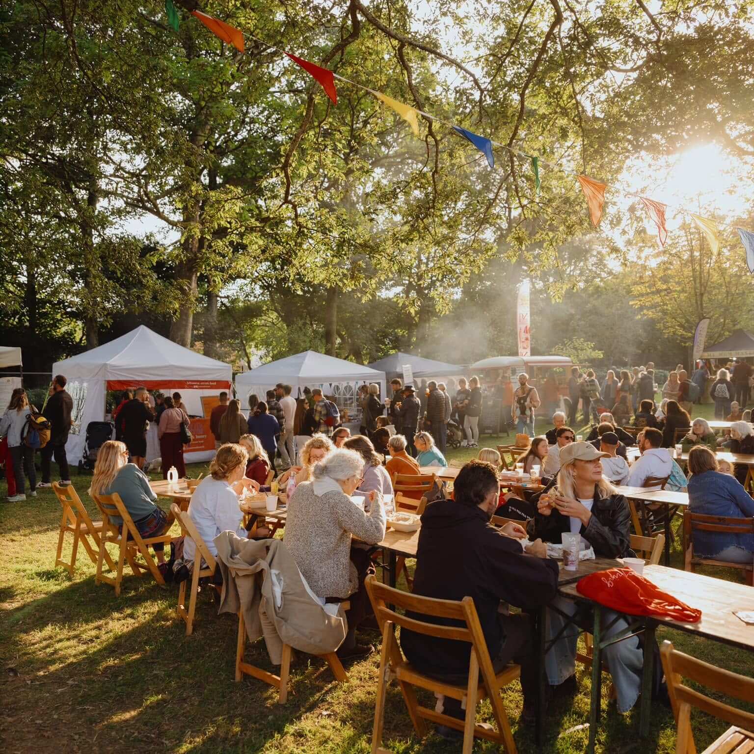 Crowds eating at long tables under trees hung with bunting at a Penzance food festival in golden evening light, with food tents and stalls behind