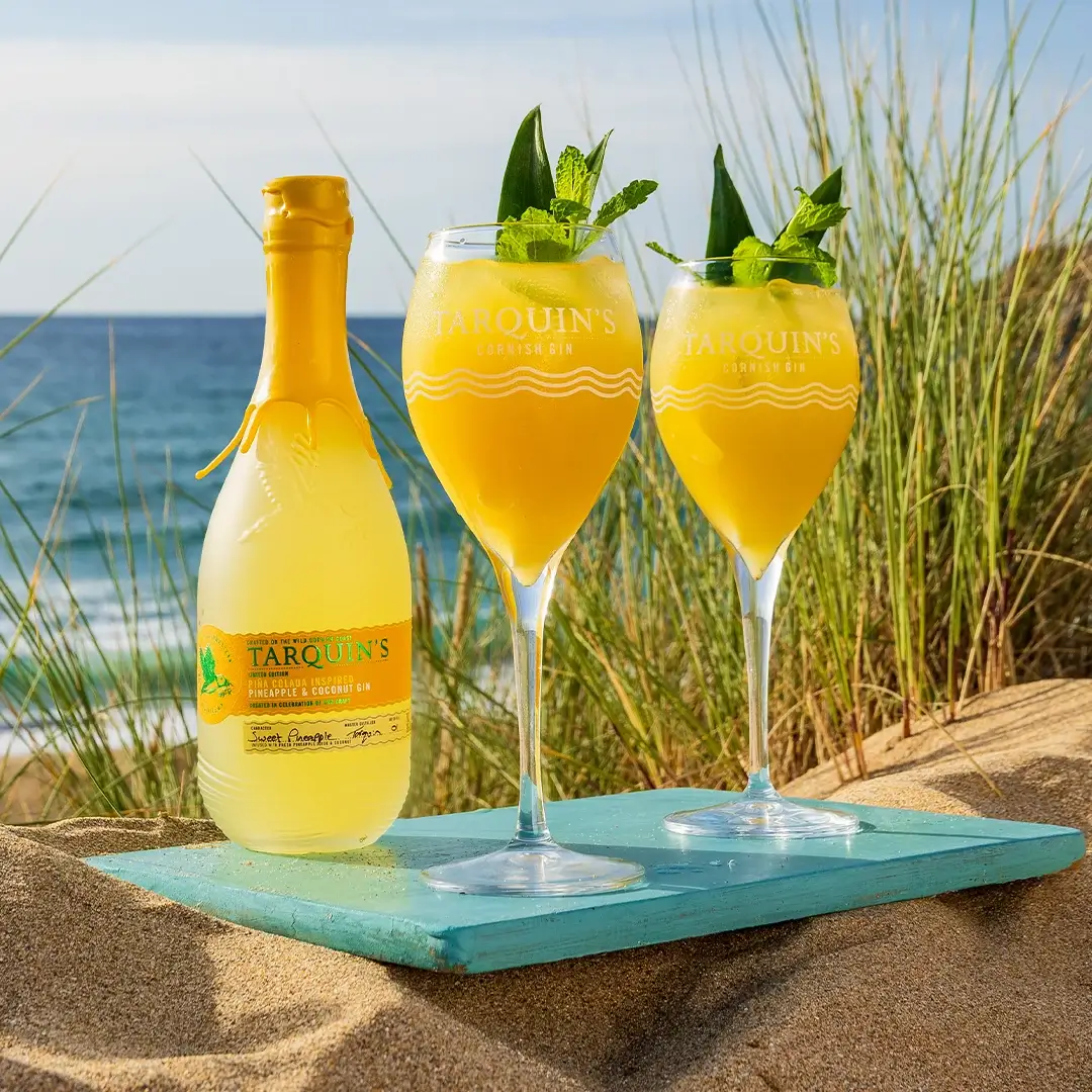 Tarquin's Piña Colada gin bottle beside two cocktails on the sand