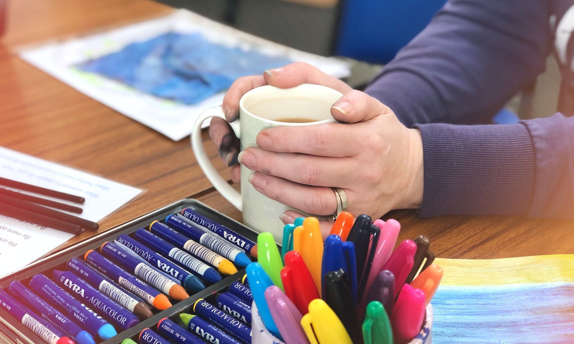 Hands around a mug of tea beside a tin of colouring crayons at a Quiet Connections creative session