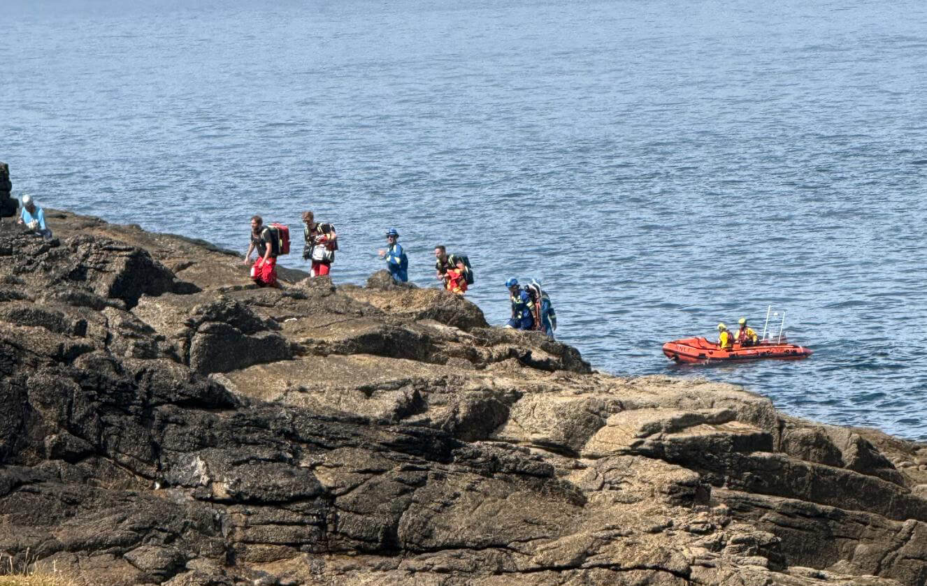 Emergency service crews walking across rocks at Pedn-men-du as an RNLI D class inshore lifeboat stands by in the water