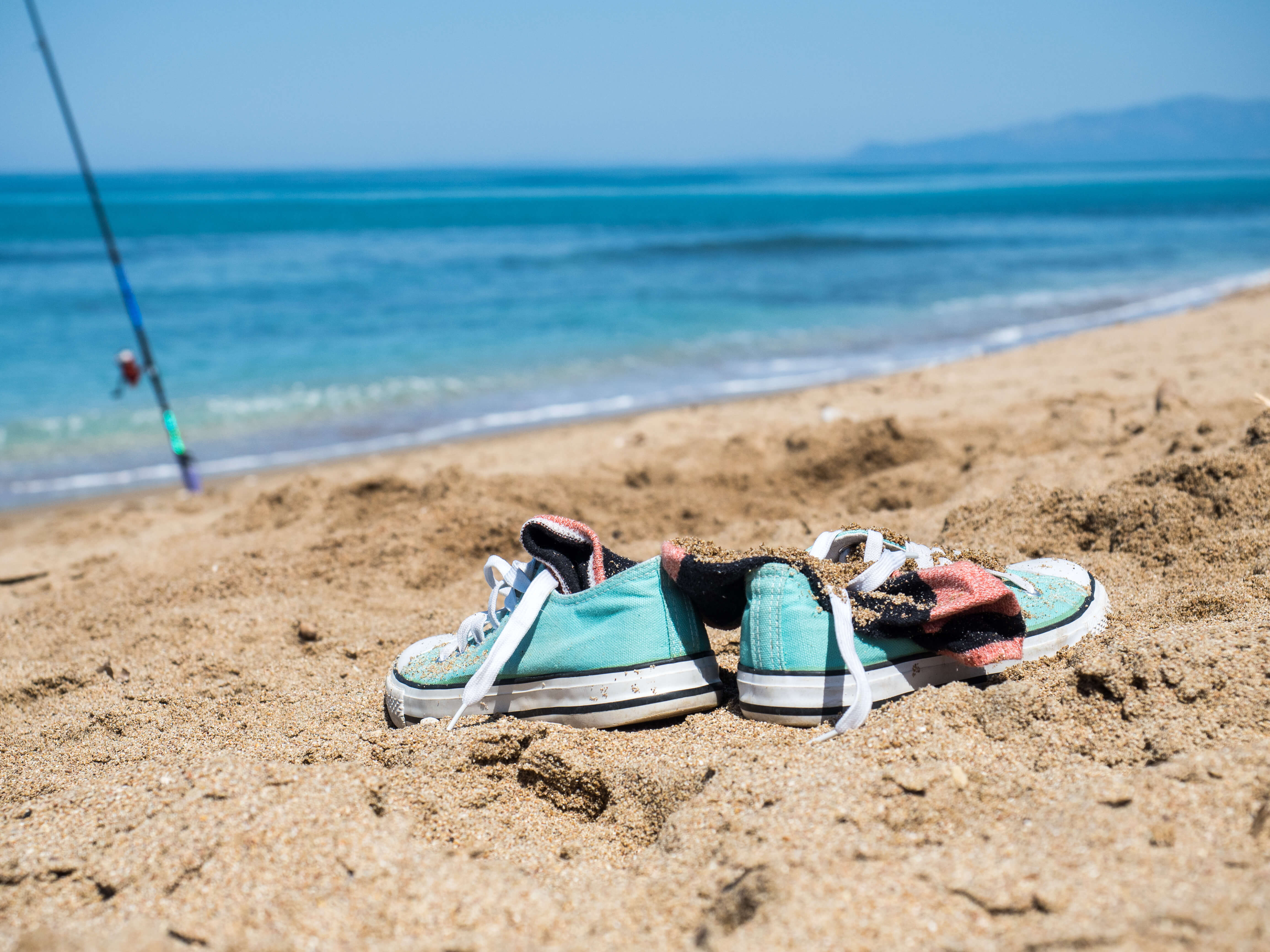 A pair of turquoise canvas trainers and a sock half buried in the sand on a Cornish-style beach, waves and a fishing rod behind