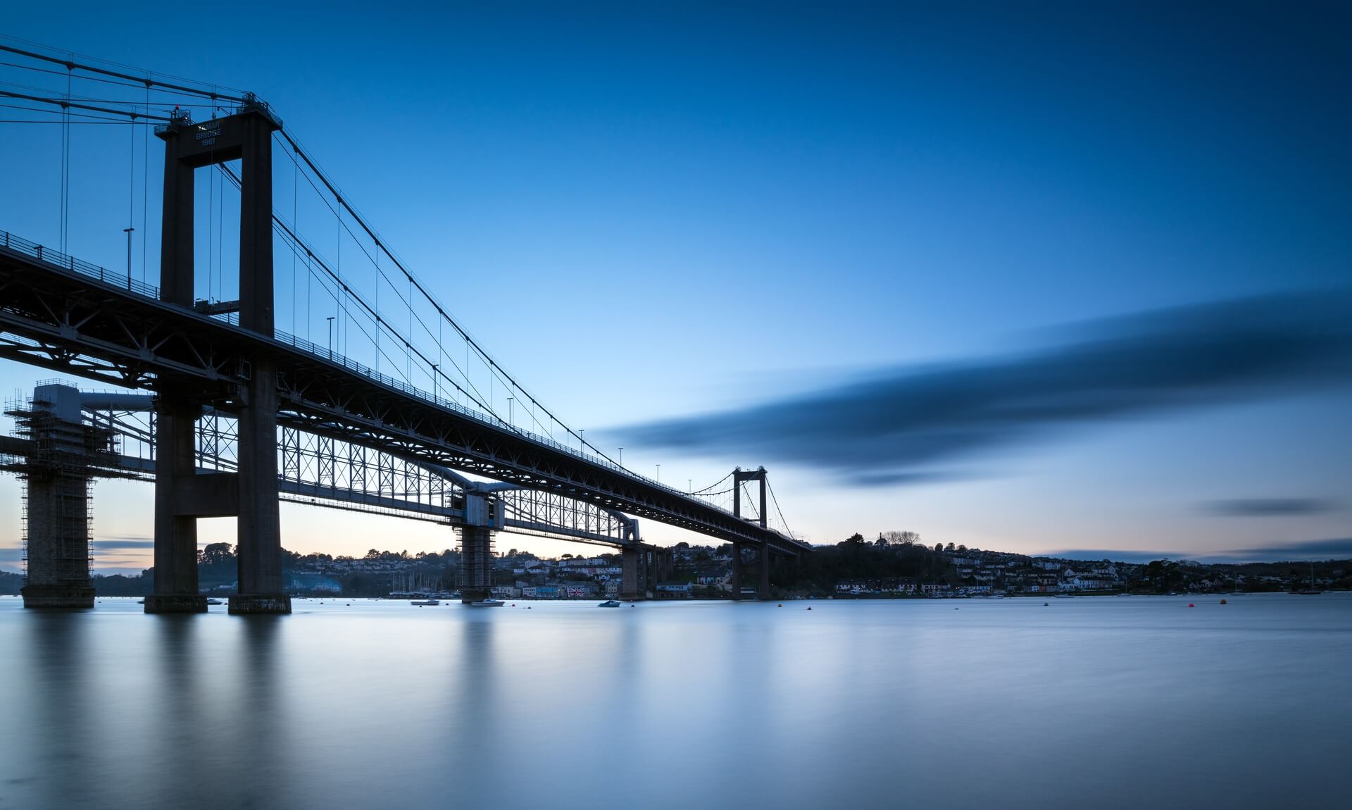 The Tamar Bridge at dusk, viewed from the water in a long-exposure shot with still reflections and a deep blue sky
