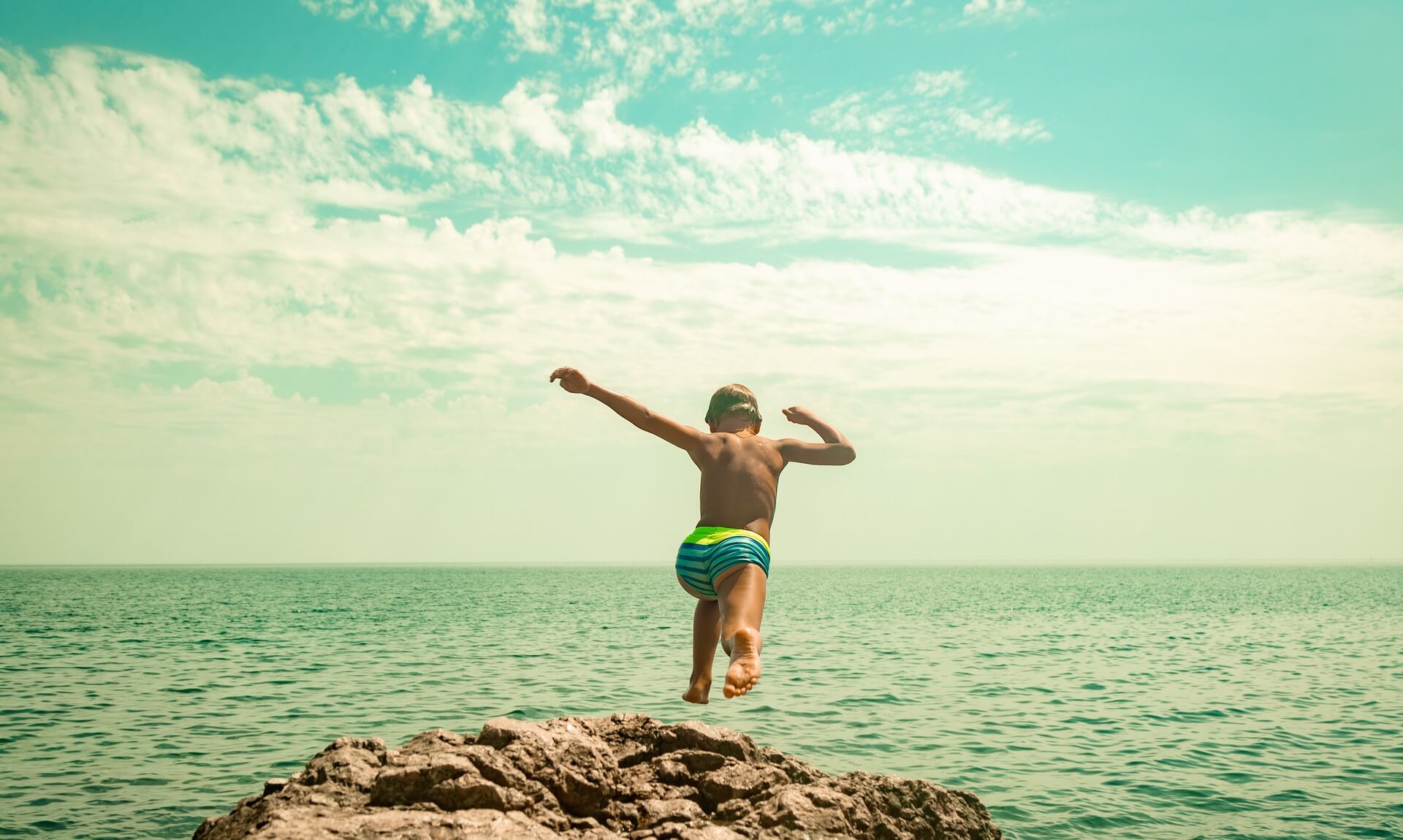 A young boy jumping feet-first from a rock into the sea