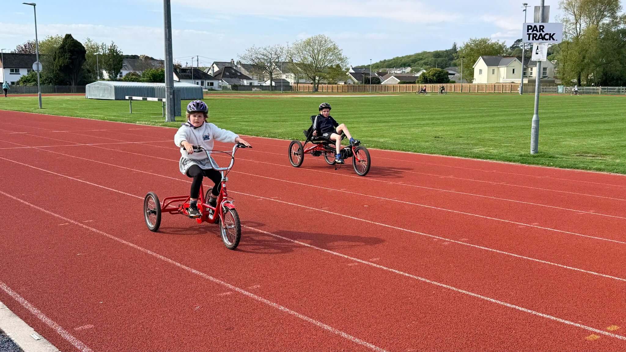 Two children on adapted cycles on the running track at Par Track, with the PAR TRACK sign and houses behind