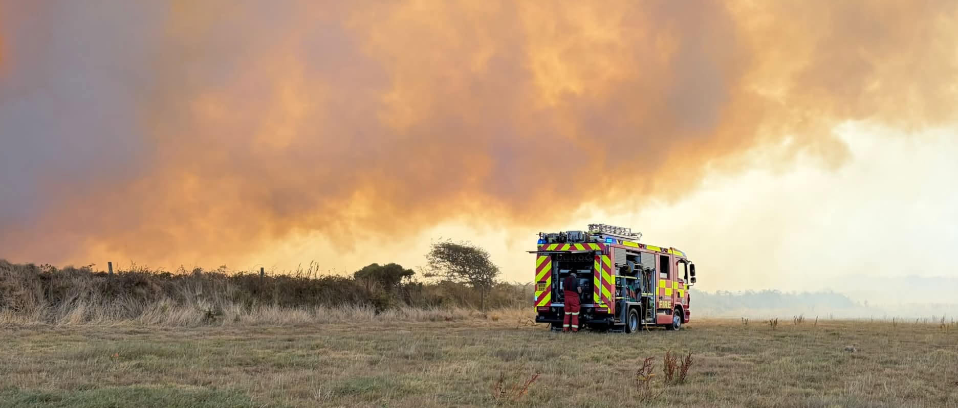 Fire engine parked in a dry field beneath a huge wall of orange wildfire smoke at Chapel Carn Brea
