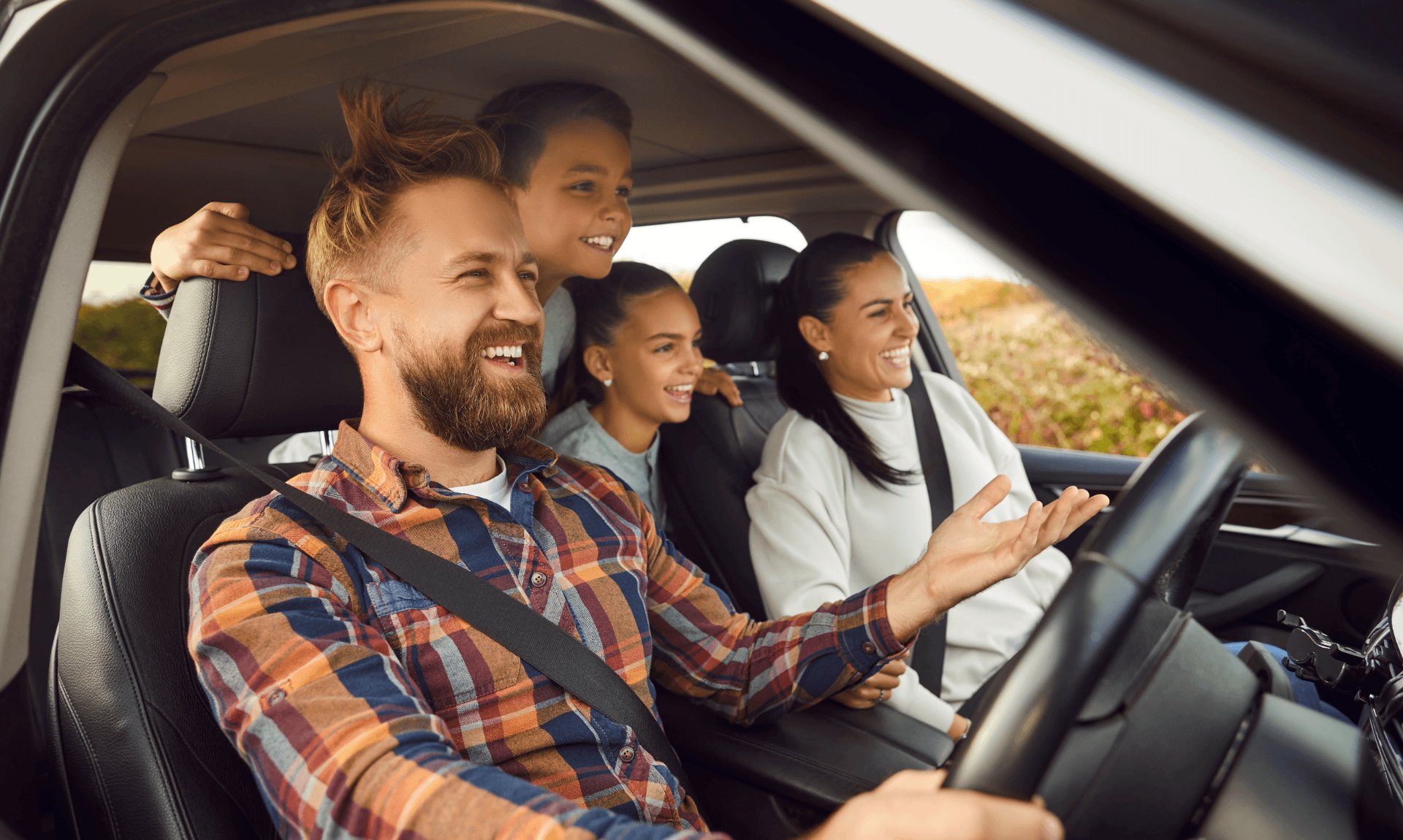 A family of four smiling in a car on a summer road trip, dad driving with a child leaning through from the back seat
