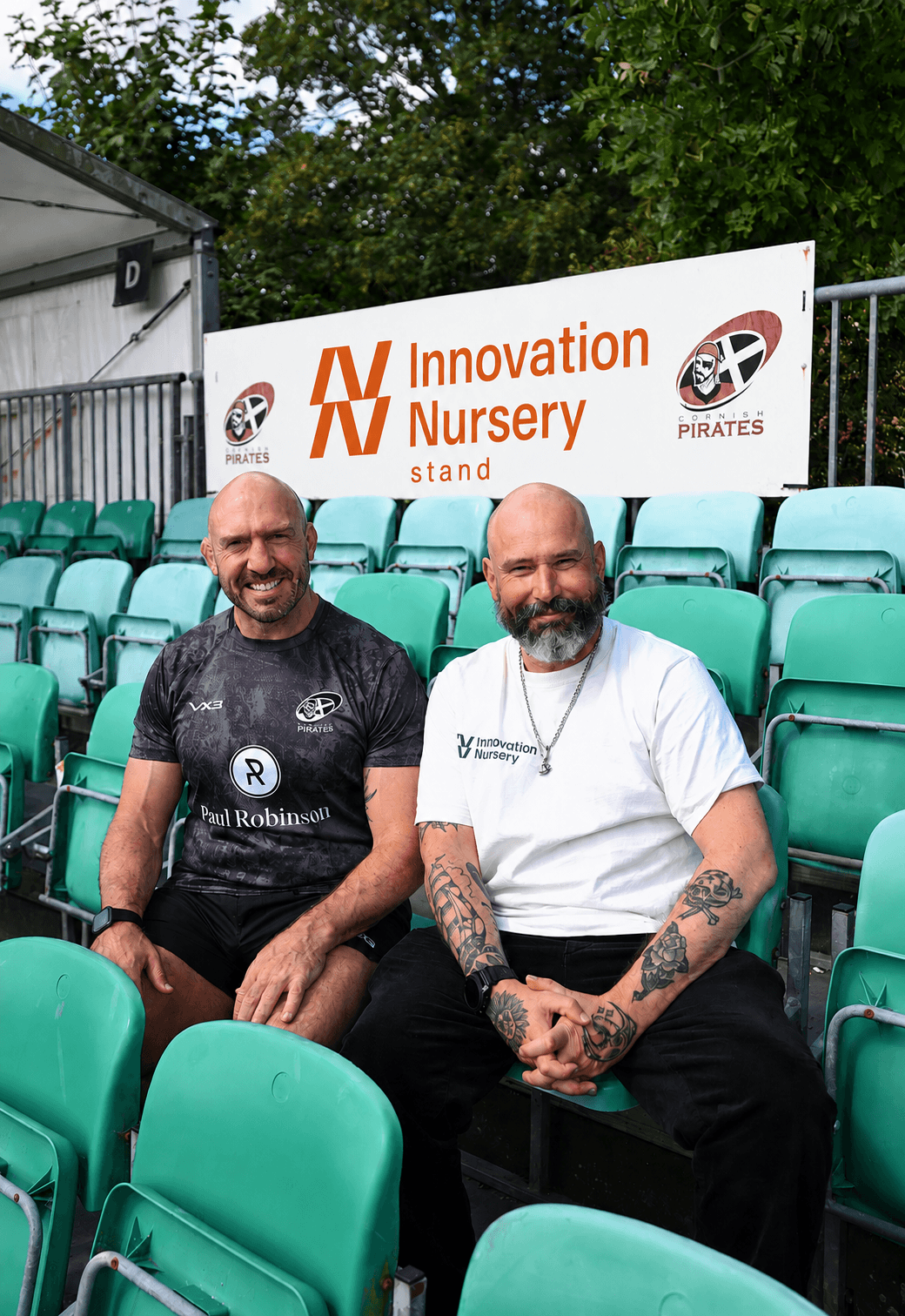 Two men sitting in stadium seating below the newly branded Innovation Nursery stand sign at the Cornish Pirates