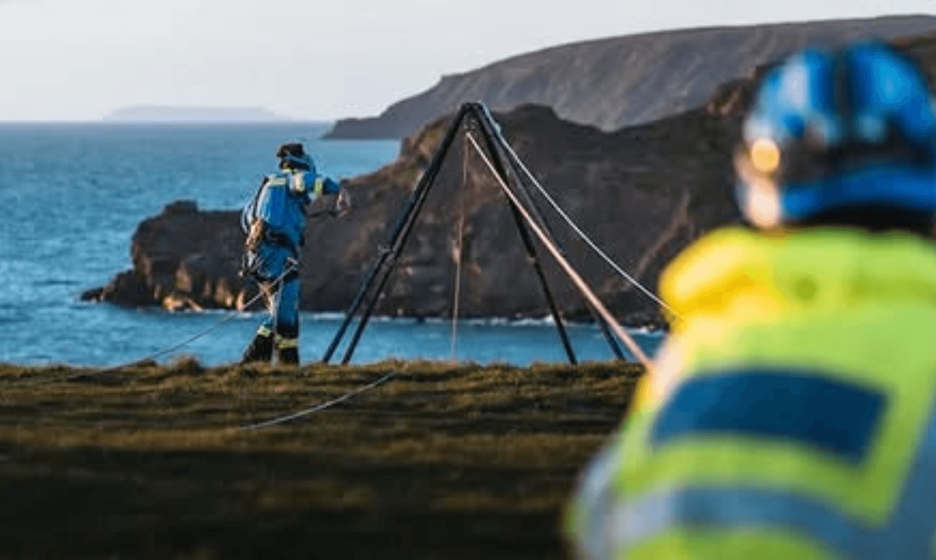 Coastguard rescue officer in helmet and harness working near a cliff edge with a rescue tripod above the sea