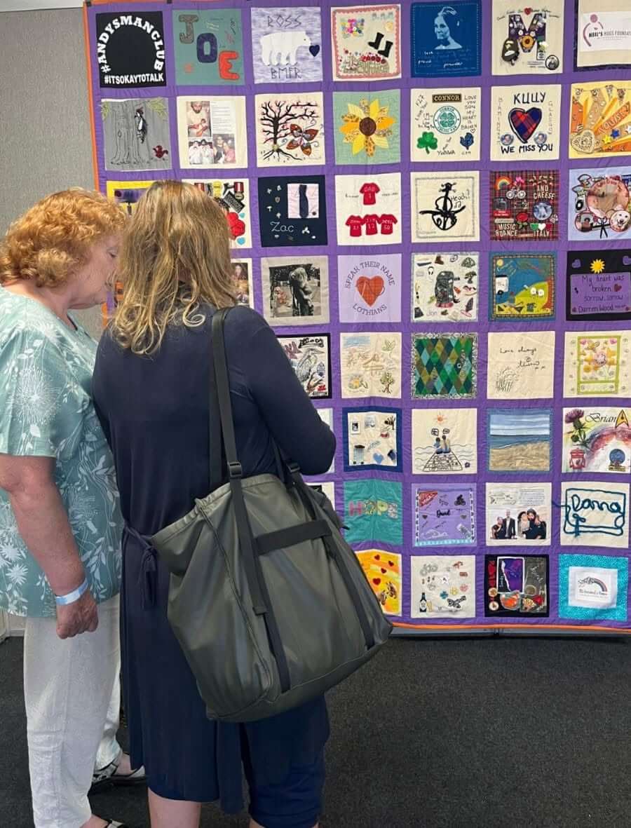 Two visitors looking at a purple-bordered Speak Their Name memorial quilt