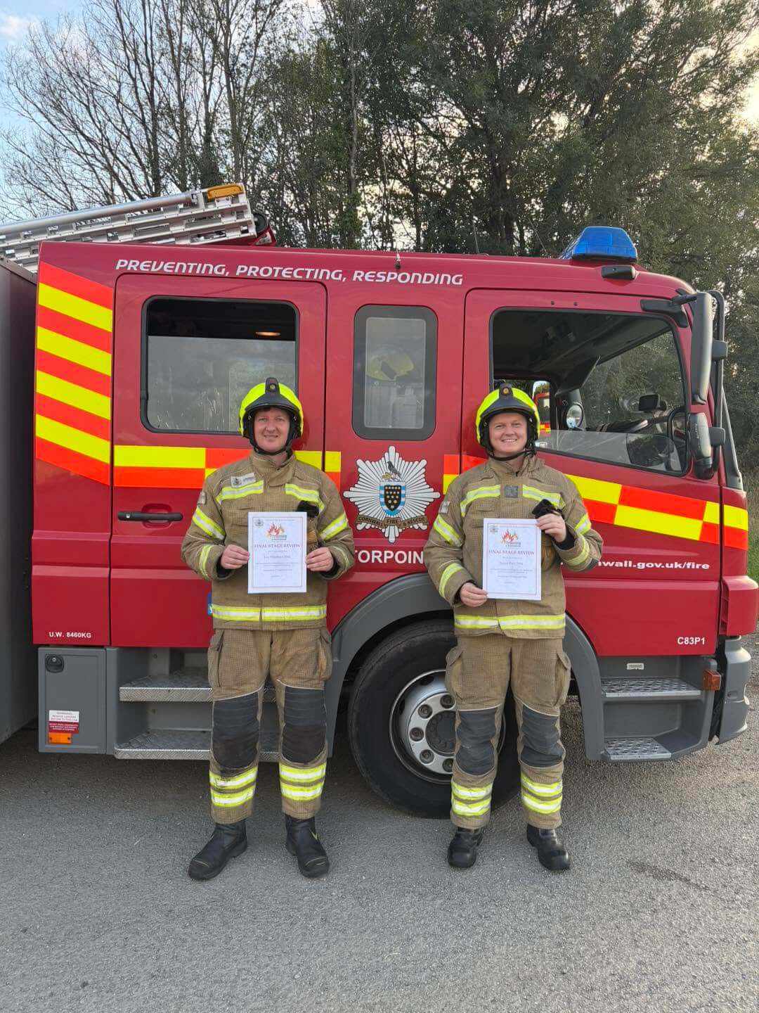 Firefighters Lee Shepherd and James Dart holding their final stage review certificates in front of a Torpoint fire engine